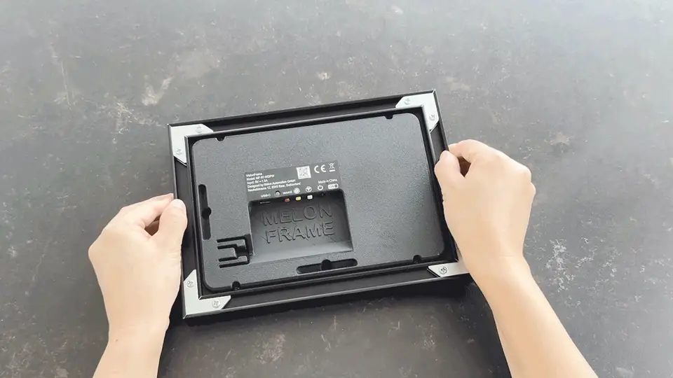 Hands moving the MelonFrame display module from a black metal frame into a solid walnut frame